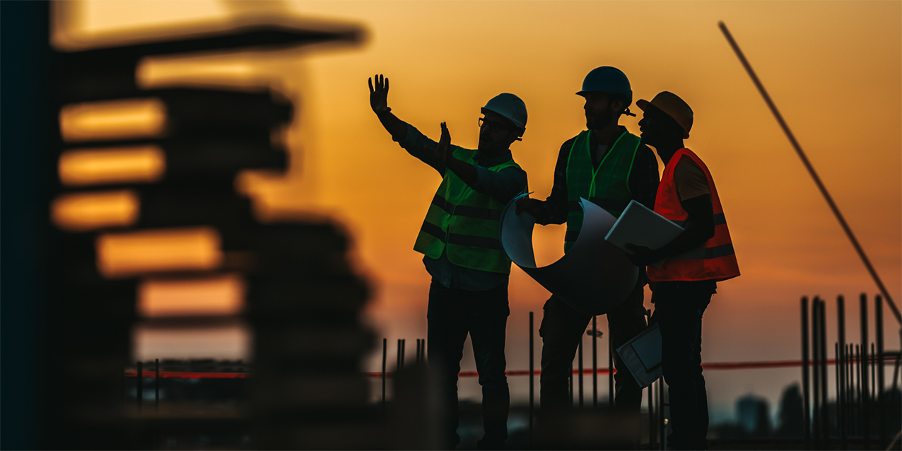 Silhouetted construction workers wearing safety gear discussing project plans at sunset.